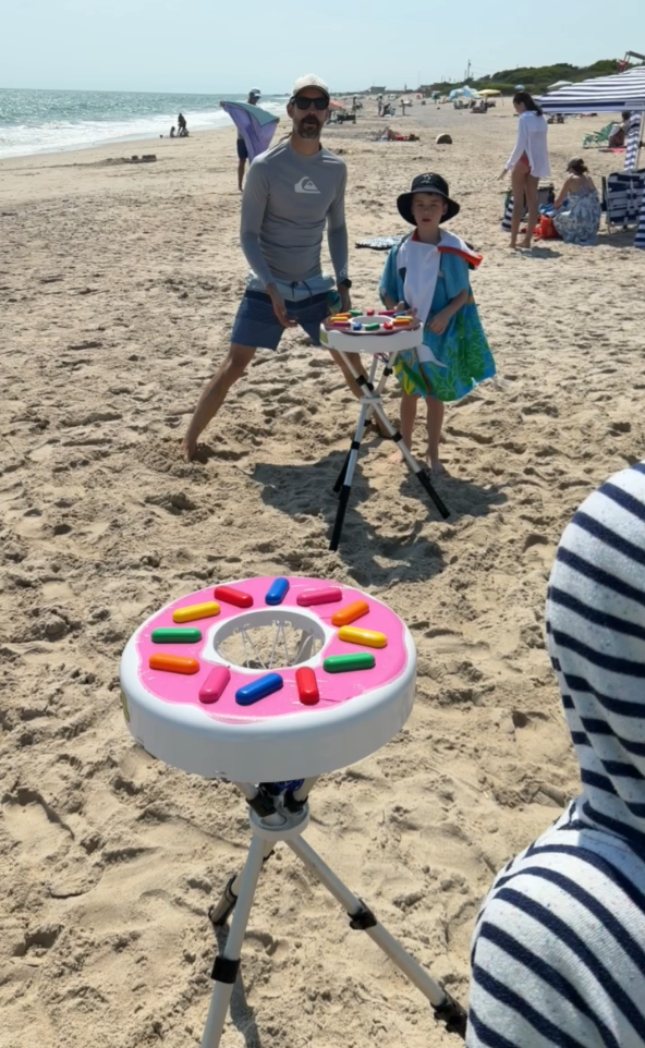 A beach scene with a man and a child standing behind a Donutball game set. In the foreground, another person in a striped hoodie is partially visible, and the beach, ocean, and other beachgoers are in the background.